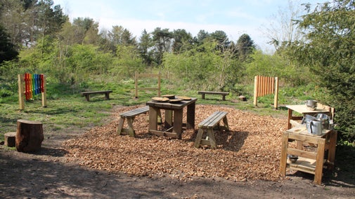 The Mud kitchen area in the woodland at Moseley Old Hall features a wooden table, benches and sinks, with metal pots and pans scattered around. There are also colourful musical instruments on stands, such as a xylophone and hanging triangles.